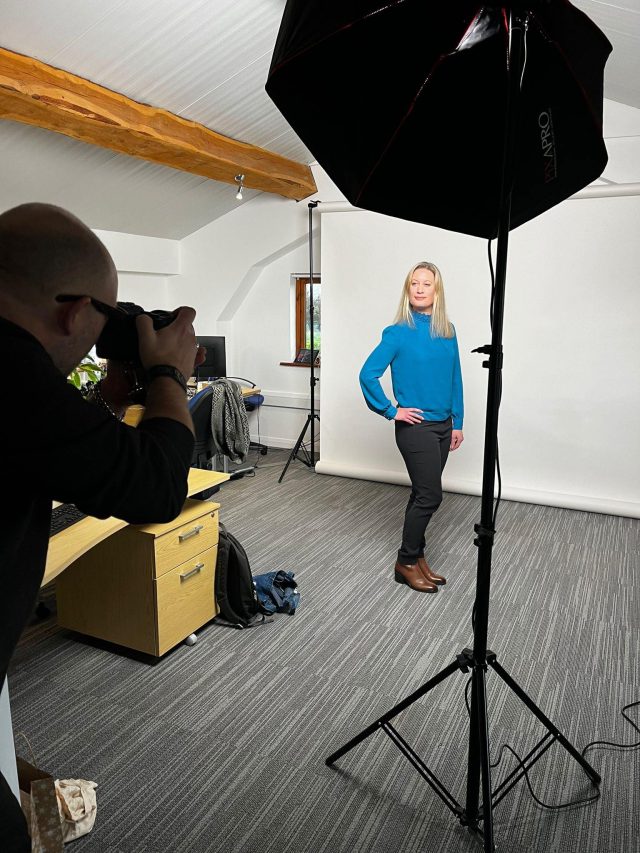 A smiling blonde professional woman in a blue blouse stands in an office which has been transformed into a photo studio, being photographed by a photographer. 