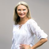 A professional woman with blonde hair wearing a white blouse with lace detailing, smiling at the camera.