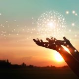 A pair of silhouetted hands lift a glowing, interconnected digital sphere against a warm sunset sky, symbolising global connectivity, innovation, and the fusion of technology with human potential.